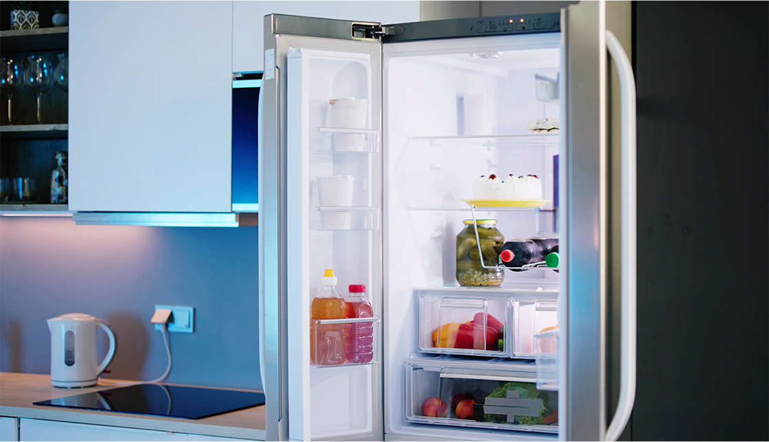 [라이프케어] 모던한 주방에 놓여 있는 양문형 냉장고의 문이 열려있고 안에는 각종 야채와 음료수, 케이크가 놓여있습니다.