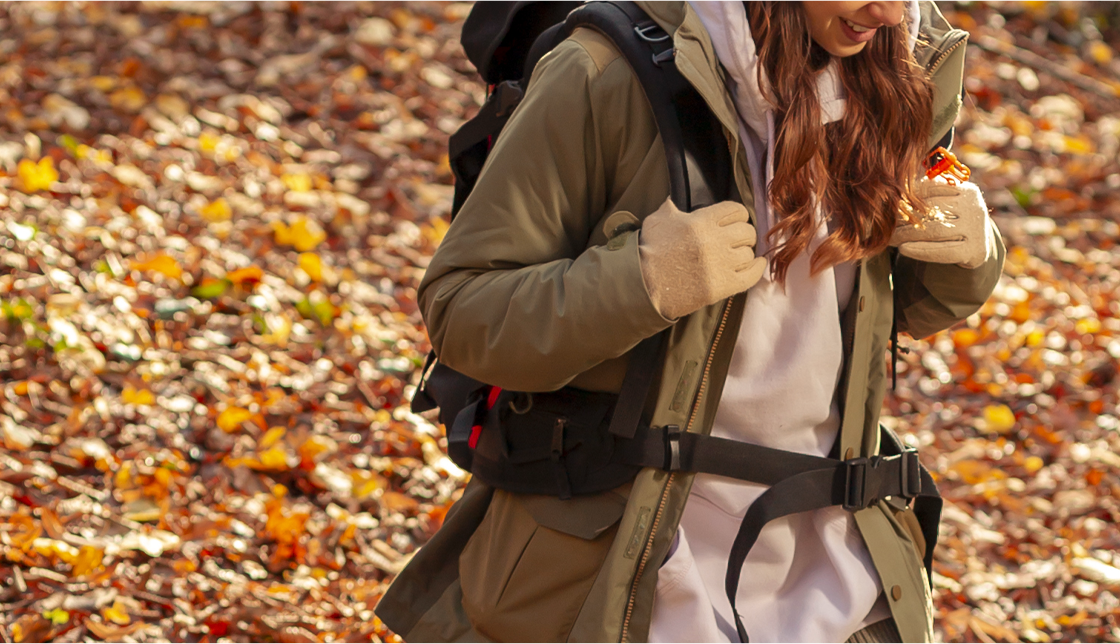 [라이프케어] 바닥에는 낙엽이 많이 떨어져 있고 머리가 길고 파마를 한 여성이 등산복을 입고 장갑을 끼고 등산가방을 메고 웃으며 등산을 하고 있습니다.