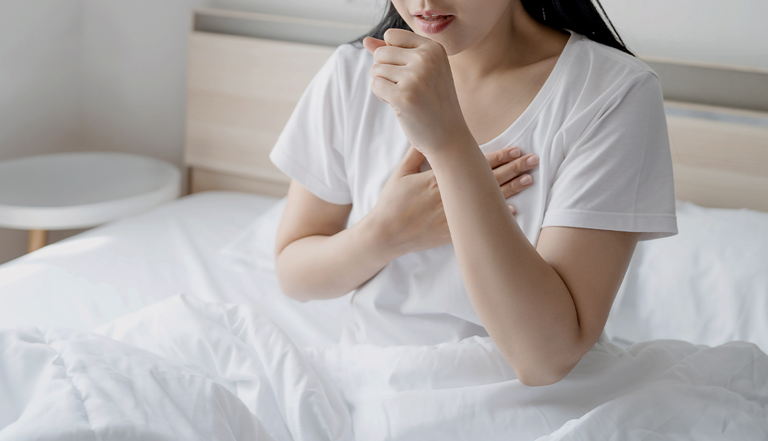 [라이프케어] 침실에서 머리가 긴 여성이 오른손은 가슴에 얹고 왼손은 주먹을 쥐어 입 앞에 두고 기침을 하는 듯한 행동을 하고 있습니다.